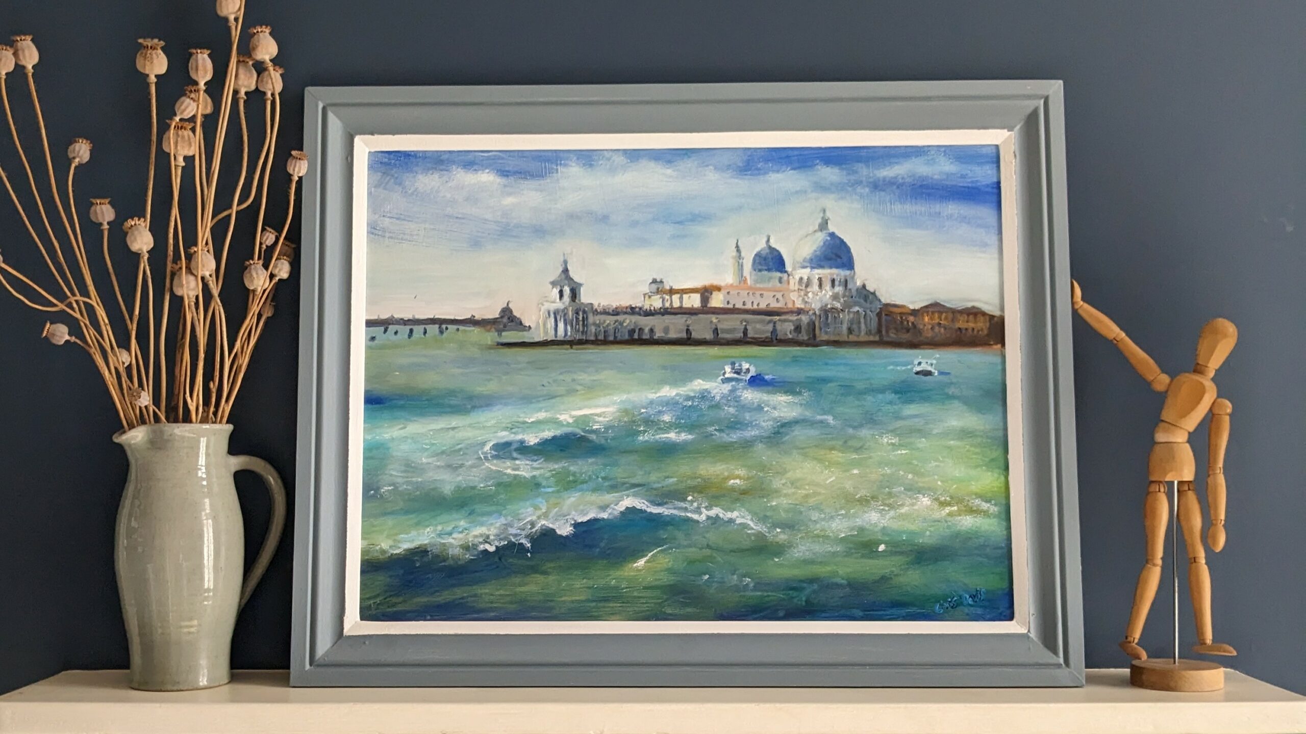A painting of Venice is propped up on a white mantelpiece; there is a blue jug on the left and a wooden mannequin on the right